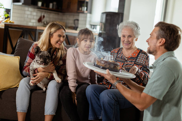 Scena commovente si svolge come una famiglia multigenerazionale si riunisce su un divano per presentare una torta di compleanno a una nonna deliziata, creando ricordi da custodire. - Foto, immagini