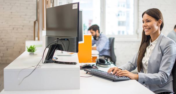 Attraente giovane dipendente donna in giacca e cravatta utilizzando il computer mentre si siede alla scrivania in ufficio. - Foto, immagini