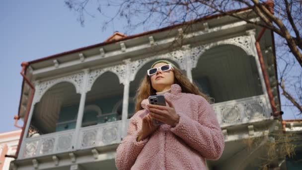 Una mujer de moda se para con un teléfono móvil en sus manos y lee noticias divertidas o se comunica en línea con amigos en la calle con gafas de sol. Una chica hipster camina con un teléfono inteligente y es - Imágenes, Vídeo