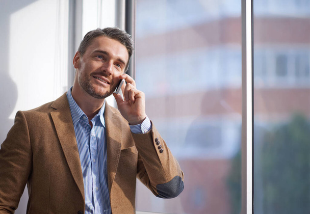 Business, phone call and man at window in office networking, deal negotiation or conversation. Communication, businessman or advisor with smartphone for consulting, connection or agenda at startup. - Photo, Image