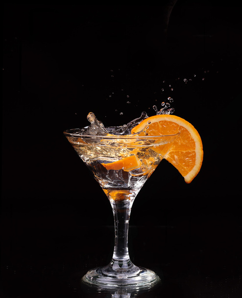 Fetta di limone spruzzata in un bicchiere d'acqua con uno spray di gocce d'acqua in movimento sospese nell'aria sopra il bicchiere su uno sfondo scuro
. - Foto, immagini