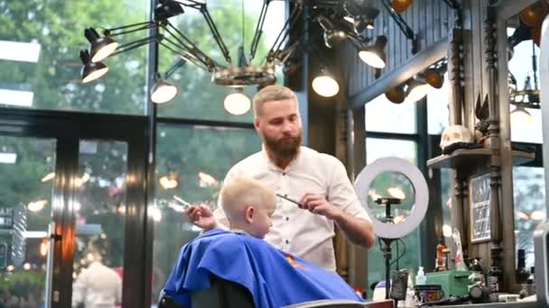A férfi fodrász fésülködéssel és ollóval vágja a kisfiú haját. Egy felnőtt férfi vágja le a gyerekét, valószínűleg borbély. Professzionális fodrász és aranyos ügyfél a modern fodrászatban. - Felvétel, videó