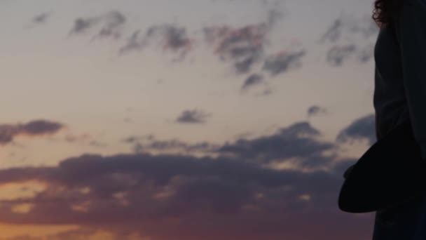 Egy női sziluett gördeszkával a kezében sétál a töltés mentén naplementekor lassított felvételben. Stílusos hipster nő visszatér haza után longboard edzés - Felvétel, videó