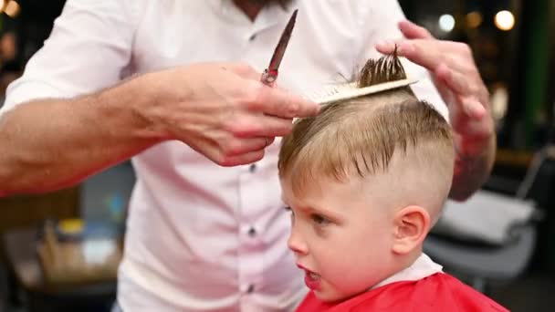A férfi fodrász fésülködéssel és ollóval vágja a kisfiú haját. Közelkép a gyerekről, ahogy levágja a haját egy felnőtt férfi, valószínűleg borbély. Professzionális fodrász és aranyos ügyfél a modern fodrászatban. - Felvétel, videó