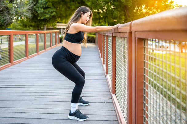Full length side view of sporty pregnant woman doing squats over fence outdoors. Pregnancy and healthy lifestyle concept. - Photo, Image