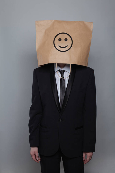 Ανώνυμος επιχειρηματίας κρύβει το κεφάλι του πίσω από χαρούμενο χαμόγελο emoticon σε γκρι φόντο - Φωτογραφία, εικόνα