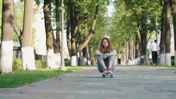 Cute carefree girl rolls down the mountain along the alley sitting on a skateboard with her arms outstretched. Walk and rest in the summer park, a young woman has fun doing active sports. - Footage, Video