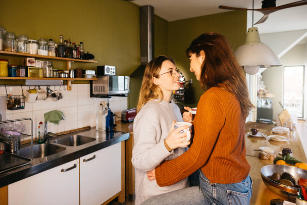 Молода пара-жінка ділиться чашкою чаю на своїй домашній кухні. Лесбійська пара насолоджується миттю разом, сіючи чай в комфорті своєї домашньої кухні. - Фото, зображення