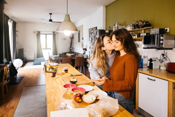 Νεαρό θηλυκό ζευγάρι φιλιά, ενώ προετοιμασία πρωινό στην κουζίνα του σπιτιού τους. Ένα ζευγάρι λεσβιών μοιράζεται μια οικεία στιγμή μαζί, απολαμβάνοντας το τσάι και την ανταλλαγή φιλιών στη ζεστή ατμόσφαιρα της κουζίνας τους στο σπίτι. - Φωτογραφία, εικόνα