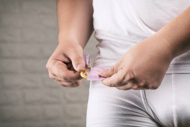 Mężczyzna w białej bieliźnie wyciąga prezerwatywę z opakowania. Metody antykoncepcji. - Zdjęcie, obraz