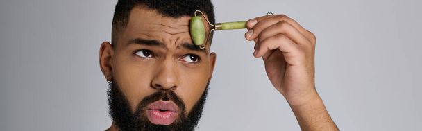African american stylish man holds face roller for skincare routine. - Photo, Image