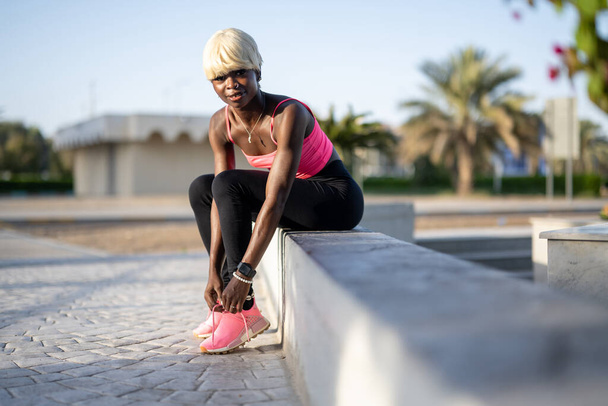 Junge Fitness-Afroamerikanerin mit blonden Haaren in Sportbekleidung bindet Schnürsenkel, während sie draußen auf Betonwänden sitzt - Foto, Bild