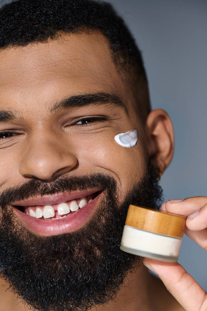 Bearded man joyfully holds jar of cream during skincare routine. - Photo, Image
