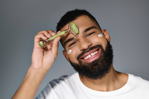 Afro-Amerikaanse man met baard met crème op zijn gezicht met behulp van gezicht roller. - Foto, afbeelding