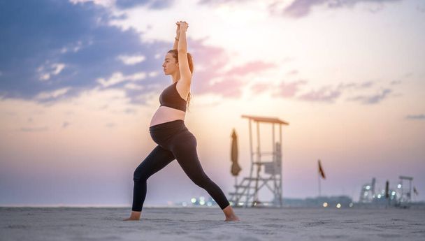 Pełna długość boczny widok portret ciężarnej kobiety w czarnej odzieży sportowej podejmowania Crescent Lunge jogi poza na plaży o zachodzie słońca. Ćwiczenia, joga i koncepcja ciąży. - Zdjęcie, obraz