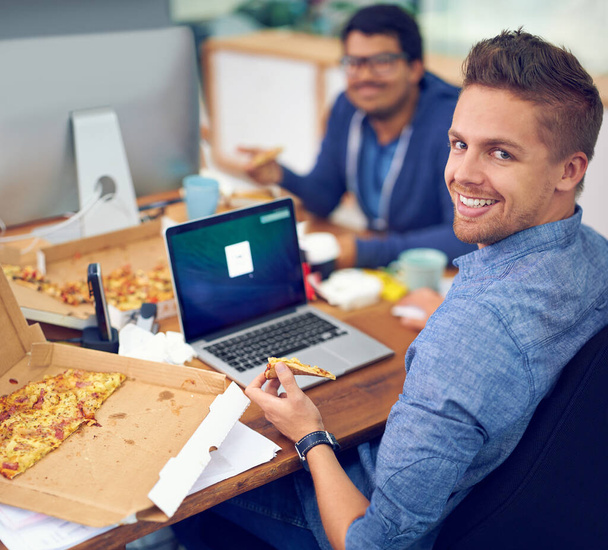 Businessman, portrait and eating by laptop in office with happiness for lunch break, pizza and coworking company. Programmer, employee and face or smile with fast food, coding team or screen at desk. - Photo, Image