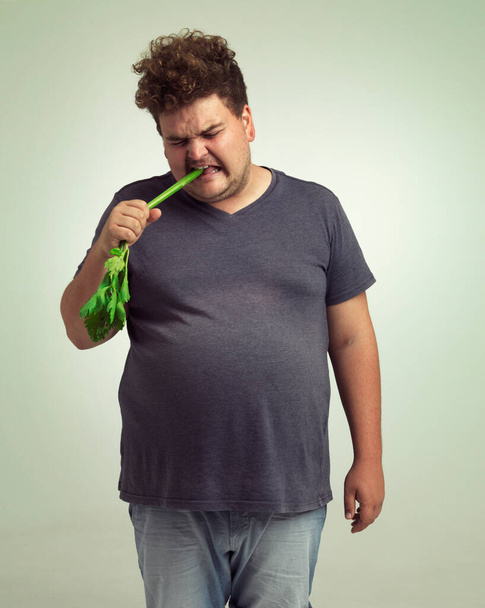 Η παχυσαρκία, η αύξηση βάρους και το χιούμορ για τον άνθρωπο στο στούντιο με σέλινο για την υγιεινή διατροφή, διατροφή και διατροφή. Υπέρβαρο άτομο με λαχανικά στο στόμα και κραυγή για πείνα, και τα τρόφιμα με βιταμίνη για κόμικ. - Φωτογραφία, εικόνα