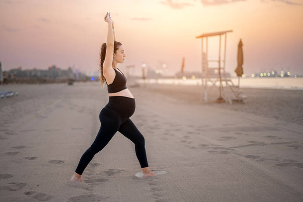 Jeune femme enceinte en tenue de sport noire faisant poser le yoga Crescent Lunge sur la plage au coucher du soleil. Entraînement, yoga et concept de grossesse. - Photo, image