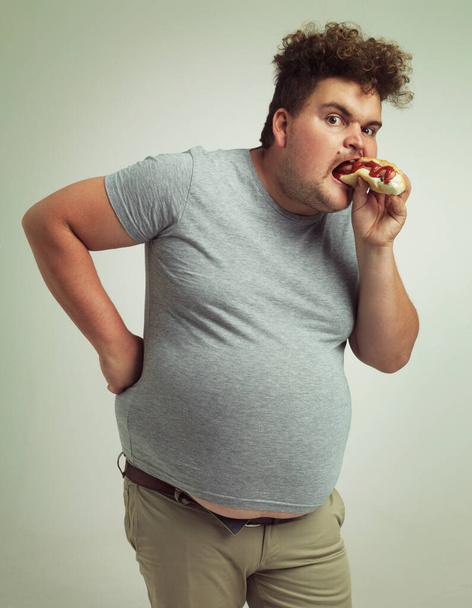 Man, fast food and eating hotdog for lunch in studio background for snack, hunger and craving for plus size guy. Male person, hungry and burger or takeaway meal for starving and enjoy while standing. - Photo, Image