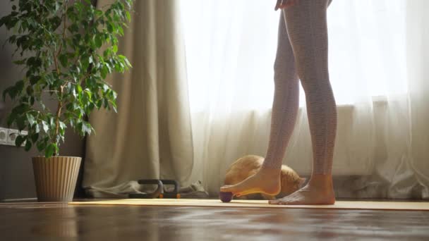 La mujer se calienta los pies con una bola de masaje en una alfombra deportiva en casa en su habitación, sus piernas en mallas deportivas de cerca. Entrenamiento deportivo en casa en un día soleado. - Imágenes, Vídeo