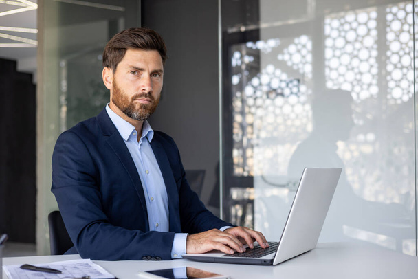 Portrait of a serious and self-confident young businessman sitting in the office at the table, typing on a laptop, working with clients and documents, looking at the camera. - 写真・画像