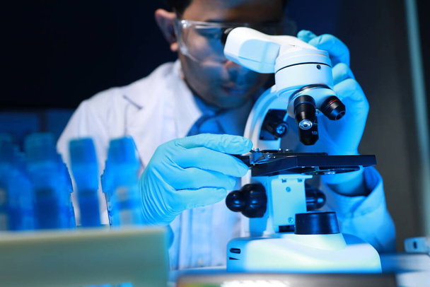 Ένας ταλαντούχος επιστήμονας κάθεται στο εργαστήριό του ενώ χρησιμοποιεί ένα μικροσκόπιο για να ψάξει για μικροσκοπικούς οργανισμούς σε ένα δείγμα δοκιμής, μικροσκόπιο που χρησιμοποιείται από τους ερευνητές για να βρουν νέα φάρμακα για τη θεραπεία των ανθρώπων. - Φωτογραφία, εικόνα