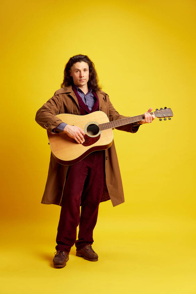 Man with long hair, musician in coat playing guitar against bright yellow background. Performer. Concept of music, performance, art, entertainment, festival, performance, ad - Φωτογραφία, εικόνα