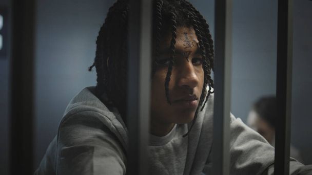 Angry African American teenage prisoner with face tattoos stands in prison cell in jail and looks at camera. Young criminal serves imprisonment term for crime. Juvenile detention center. Portrait. - Photo, Image
