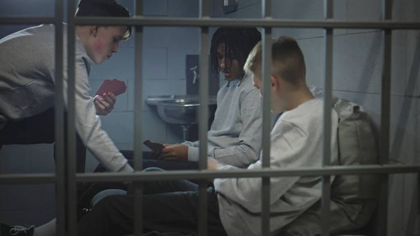 Tres diversos adolescentes juegan juegos de cartas en la celda de la prisión. Jóvenes prisioneros, criminales cumplen pena de prisión por crímenes en la cárcel. Centro de detención juvenil o correccional. Vista a través de barras de metal. - Foto, Imagen