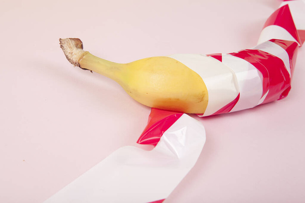 een rijpe banaan, gewikkeld in een rood en wit plastic lint, gevangen gehouden. Levendige kleur en minimale pop art fotografie - Foto, afbeelding
