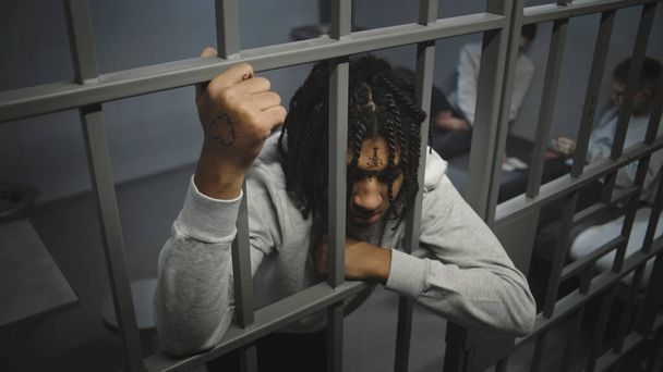 Angry African American teenage prisoner stands in prison cell in jail, holds metal bars. Young inmates play cards on bed in the background. Youth detention center or correctional facility. High angle. - Photo, Image