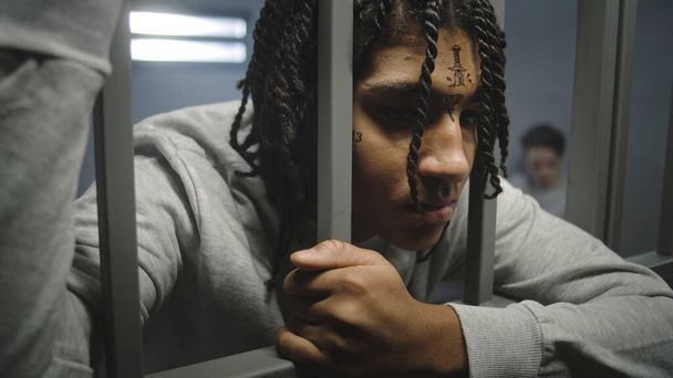 Zbliżenie zdjęcia wściekłego Afroamerykanina, nastoletniego więźnia z tatuażami na twarzy, stojącego w celi, trzymającego metalowe pręty. Młody więzień odsiaduje karę więzienia za przestępstwo w więzieniu. Ośrodek zatrzymań młodzieży. - Zdjęcie, obraz