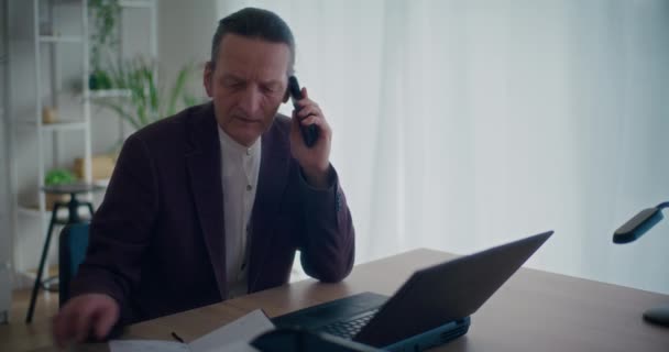 Gerente masculino mayor hablando en el teléfono móvil y escribiendo en el bloc de notas mientras trabaja en el portátil en el escritorio en la oficina - Imágenes, Vídeo