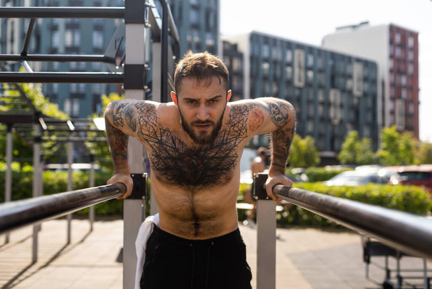 Muscular culturista guapo hombre con torso desnudo entrena tríceps en una máquina de ejercicio de cable cruzado en la calle. - Foto, imagen