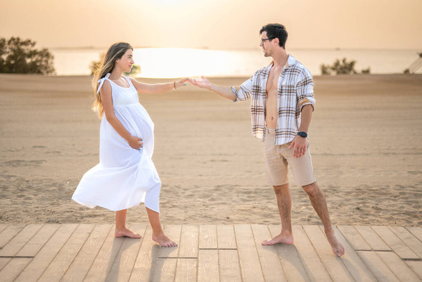 El amor de la mujer embarazada y el hombre guapo en camisa desabotonada alcanzándose las manos en la playa - Foto, Imagen
