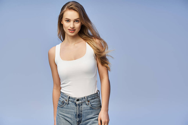 A young blonde woman exudes timeless beauty in a studio wearing a white tank top and jeans. - Photo, Image