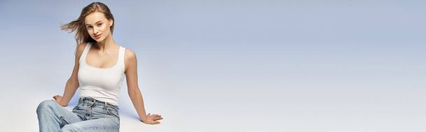 Μια νεαρή ξανθιά γυναίκα ποζάρει κομψά με λευκό πουκάμισο και τζιν σε ένα στούντιο, αποπνέοντας μια λαμπερή και αιθέρια ομορφιά. - Φωτογραφία, εικόνα