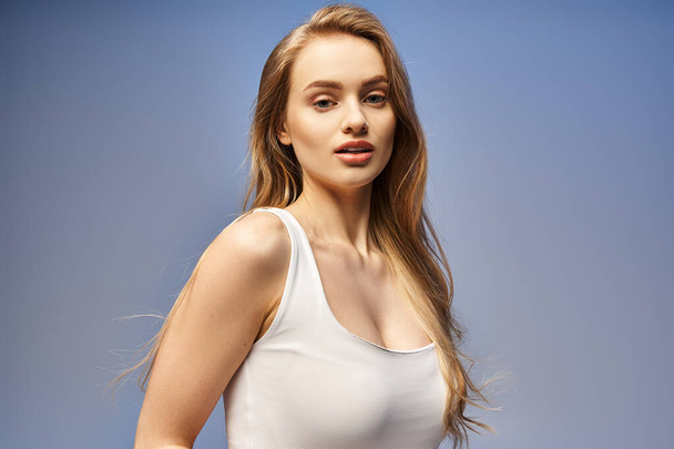 Uma jovem loira com um sorriso radiante posa graciosamente em uma blusa branca, exalando beleza e confiança em um estúdio. - Foto, Imagem