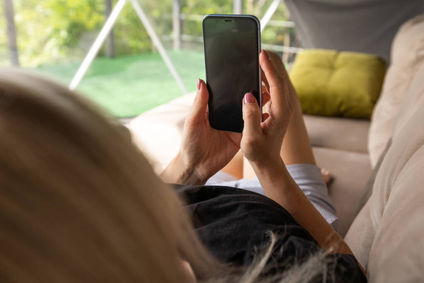Ευτυχισμένη νεαρή καυκάσια γυναίκα χαλαρώσετε σε άνετο καναπέ στο σπίτι γραπτών μηνυμάτων στο smartphone, χρησιμοποιήστε το κινητό τηλέφωνο, περιηγηθείτε ασύρματο internet στο gadget, ψώνια σε απευθείας σύνδεση από το σπίτι. - Φωτογραφία, εικόνα
