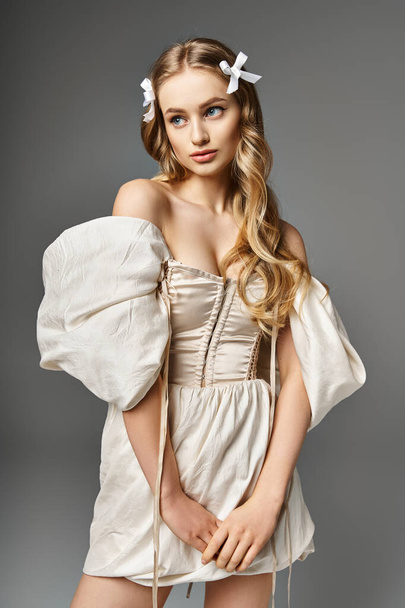 Μια νεαρή, ξανθιά γυναίκα στέκεται σε ένα στούντιο φορώντας ένα κοντό φόρεμα και ένα φιόγκο στα μαλλιά της, αποπνέοντας μια αίσθηση κομψότητας και γλυκύτητας.. - Φωτογραφία, εικόνα