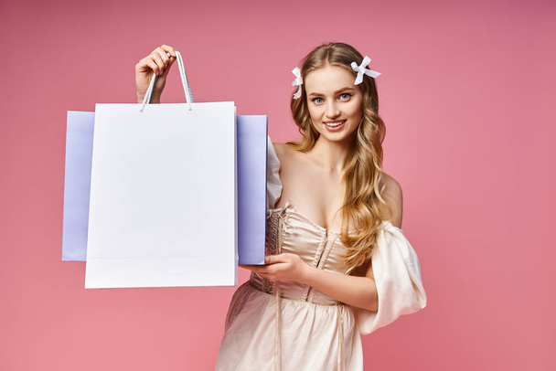 Μια εκθαμβωτική νεαρή γυναίκα με ξανθά μαλλιά με χαρά κρατά μια τσάντα για ψώνια σε ένα σκηνικό στούντιο. - Φωτογραφία, εικόνα