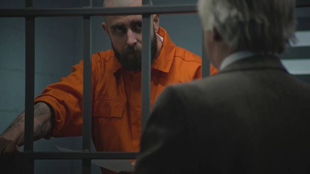 Заключенный в оранжевой форме опирается на тюремные камеры, разговаривает с адвокатом и читает адвокатский контракт. Заключенный отбывает тюремный срок за преступление в исправительном учреждении. Гангстер в СИЗО. - Фото, изображение