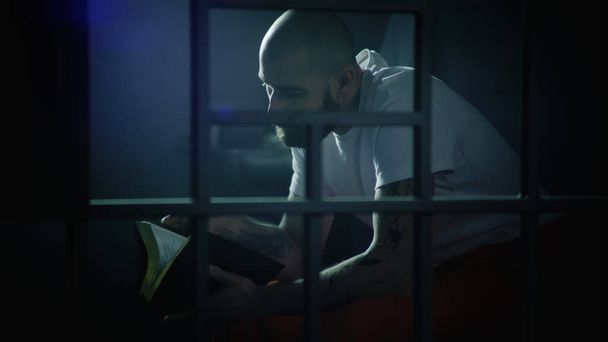 Mannelijke gevangene in oranje uniform zit op bed in de gevangenis cel, leest de Bijbel. Illegaal veroordeelde man zit gevangenisstraf uit. Zicht door metalen staven. Detentiecentrum of correctionele inrichting. - Foto, afbeelding