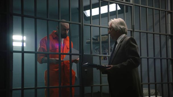 Criminel en uniforme orange se tient derrière les barreaux dans la cellule de la prison, parle avec un avocat et lit contrat. Le prisonnier purge une peine d'emprisonnement pour crime dans un établissement correctionnel. Gangster en centre de détention. - Photo, image