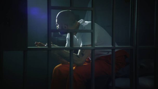 Злочинець в помаранчевій формі сидить на ліжку в камері в'язниці, встає і дивиться на заборонене вікно. В'язень відбуває термін позбавлення волі за злочин у в'язниці. Гангстер у СІЗО. Перегляд через металеві панелі. - Фото, зображення
