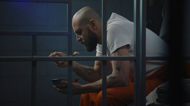 Mannelijke gevangene in oranje uniform zit op bed in de gevangenis cel, eet walgelijk gevangenisvoedsel uit ijzeren kom. Gevangene zit gevangenisstraf uit voor misdaad in de gevangenis. Detentiecentrum of correctionele inrichting. - Foto, afbeelding