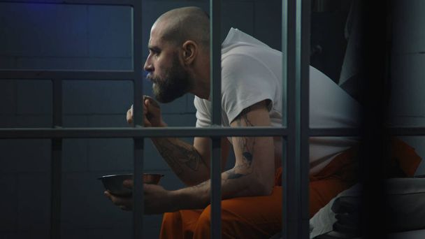 Männlicher Häftling in orangefarbener Uniform sitzt auf dem Bett in der Gefängniszelle und isst ekelhaftes Gefängnisessen aus eiserner Schüssel. Häftling verbüßt Haftstrafe für Verbrechen im Gefängnis. Haftanstalt oder Justizvollzugsanstalt. - Foto, Bild