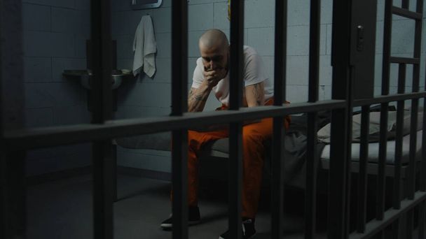 Mannelijke gevangene in oranje uniform zit op bed in de cel en probeert walgelijk gevangenisvoedsel te eten uit een ijzeren kom. Crimineel zit gevangenisstraf uit. Detentiecentrum of correctionele inrichting. - Foto, afbeelding