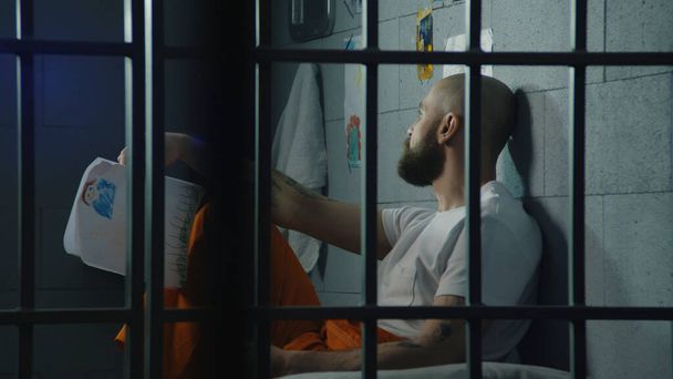 Depressieve mannelijke gevangene houdt tekeningen van het kind en kijkt naar het raam zittend op bed in de cel. Illegaal veroordeelde man zit gevangenisstraf uit. Detentiecentrum of correctionele inrichting. - Foto, afbeelding