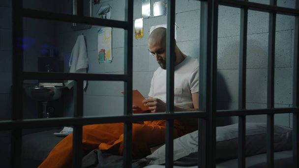 Male prisoner in orange uniform sits on bed in prison cell, looks at photographs of family and children. Criminal serves imprisonment term for crime in jail. Detention center or correctional facility. - Photo, Image